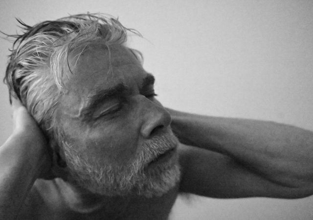 A black-and-white portrait of an older man with his eyes closed, resting his head on his hand, conveying a sense of contemplation and tranquility.