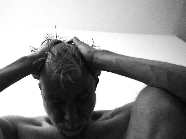 A black-and-white photograph of a person from the front, with their arms wrapped around their head, conveying a sense of introspection, comfort, and vulnerability. The image captures the raw emotion and texture of the moment, emphasizing solitude and self-reflection.