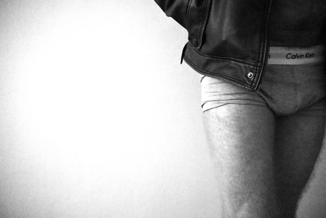 A black-and-white photograph of a person wearing a leather jacket and underwear, with the jacket slightly lifted to reveal the waistband. The image captures a moment of subtle vulnerability and contrast, emphasizing the interplay between protection and exposure.