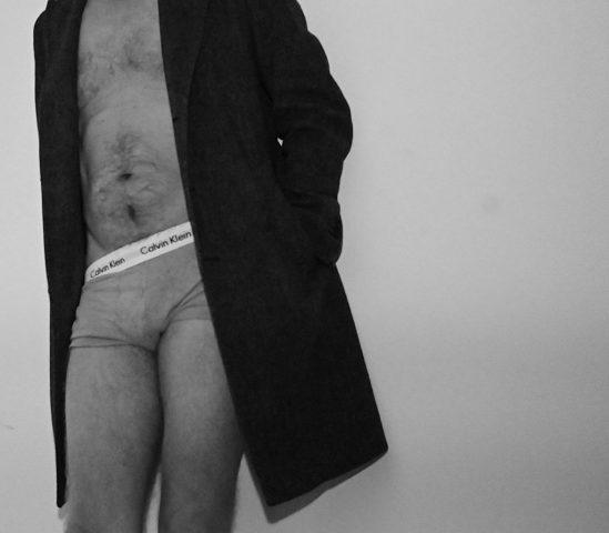 A black-and-white photograph of a person wearing a coat and underwear, emphasizing the contrast between formal attire and exposed skin. The image explores themes of vulnerability, duality, and the complexity of identity.