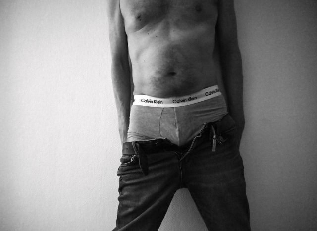 A black-and-white photograph of a person with their torso and hands in their pockets, wearing briefs and jeans. The image captures a moment of casual vulnerability, emphasizing the contrast between exposed skin and clothing, and evoking themes of identity and authenticity.